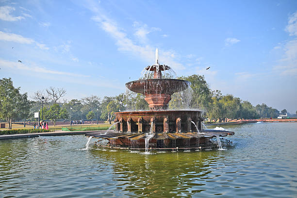 A beautiful multi-tiered fountain with water flowing gracefully, surrounded by lush greenery and clear blue skies, creating a serene park atmosphere.