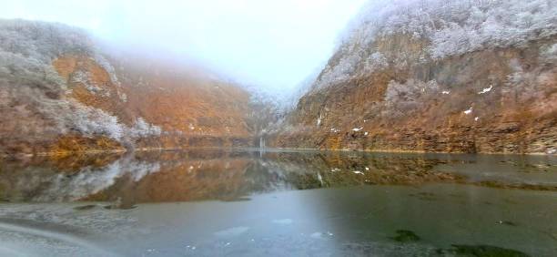 A breathtaking view of a fog-covered quarry lake, surrounded by rugged cliffs and frost-laden trees. The still, partially frozen water creates a perfect reflection of the icy slopes, enhancing the serene and mystical ambiance. The misty sky blends seamlessly with the snow-covered peaks, giving the scene a dreamlike quality. A true winter wonderland frozen in time.