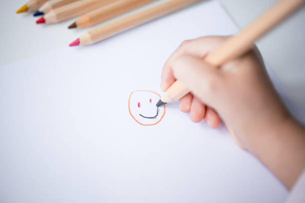 Child drawing smiley face with colored pencils on white paper.
