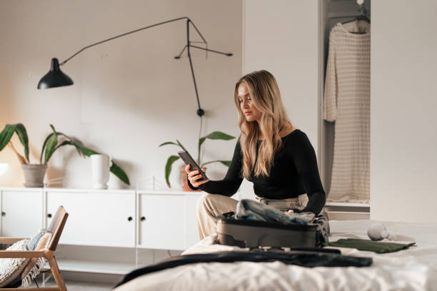 istock Woman packing clothes on bed while using a smartphone in minimalistic bedroom 2198919704