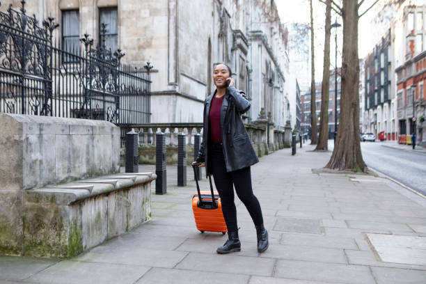 Female visitor to London wheeling an orange-coloured suitcase on a pavement whilst talking on her smartphone