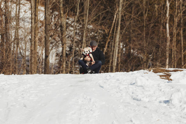 A man sitting on a toboggan at the top of a hill, holding his french english bulldog puppy, reading to slide down.