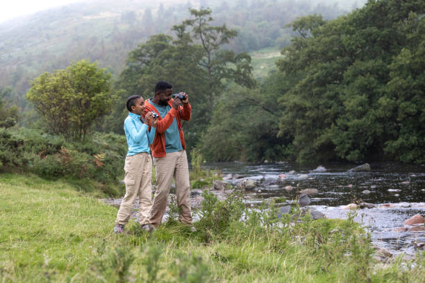 Wide full-length side view of a mid-adult black father and his young multi-ethnic daughter taking a break during a scenic countryside walk on a misty day in Ingram Valley, North East of England. They have stopped by River Breamish to take a closer look using binoculars while standing side by side. The serene landscape, tall grass, and foggy atmosphere create a peaceful and refreshing outdoor experience. Videos are available similar to this scenario.
