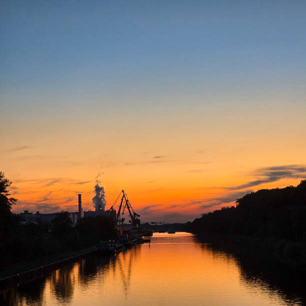 Beautiful sunset creates a serene reflection on the river, framed by silhouettes of trees and industry. Hannover Mittellandkanal