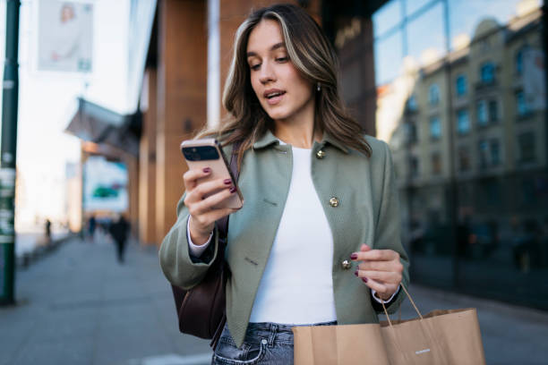 istock Young woman using smartphone while carrying shopping bag in city 2198943940