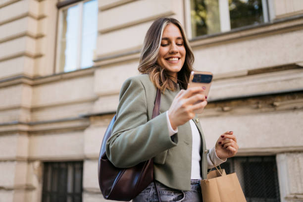 istock Smiling young woman using smartphone while carrying shopping bag in the city 2198944507