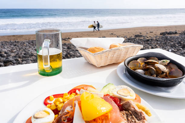 istock Seaside lunch featuring cold beer, fresh salad, steamed clams with surfers strolling along sandy coastline 2198950771