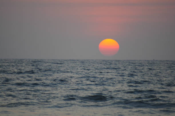 The image shows a serene ocean scene at sunset with the sun partially obscured by clouds, casting a golden reflection on the water. The calm waves and warm colors create a peaceful atmosphere.