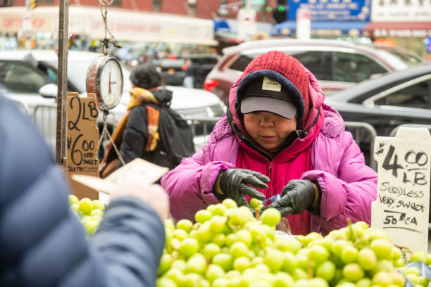 February 8th, 2025 - New York City, NY, USA: An older woman dressed in warm clothes sells grapes to customers along Canal Street in Chinatown.