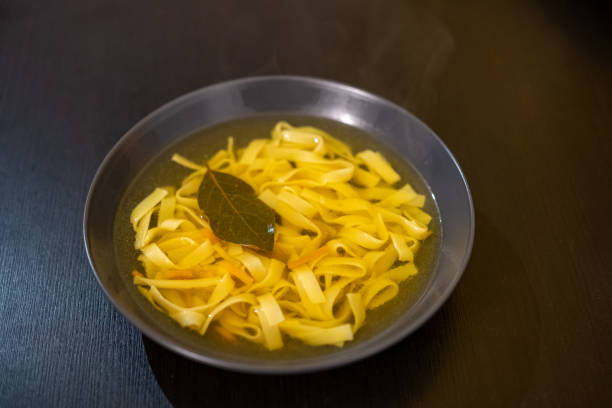 Close-up shot of a bowl of homemade chicken noodle soup with flat pasta, a bay leaf, and carrot strips in clear broth. The dish is served in a black bowl on a dark wooden table.