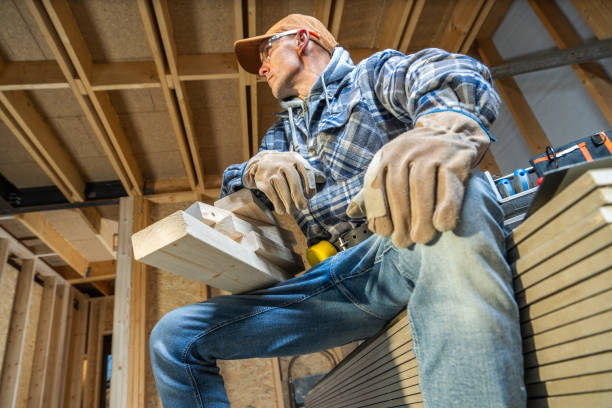 A skilled carpenter pauses to rest, seated on a stack of wood planks, while working on a project in a bright, home workshop, surrounded by tools and materials.