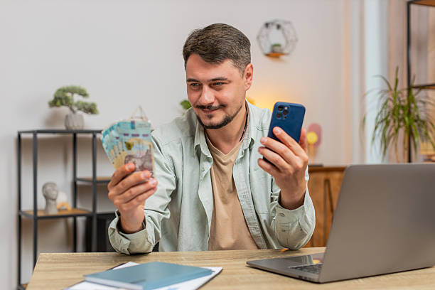 istock Smiling Caucasian young man counting money cash using smartphone at home office table in living room 2198990137