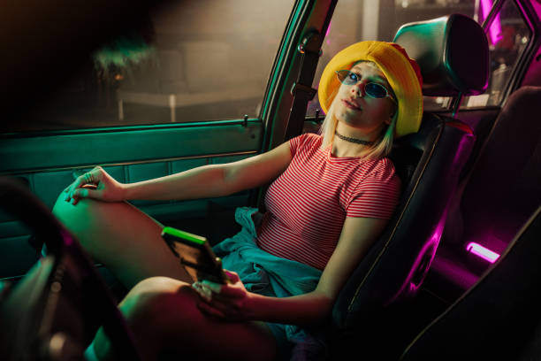 A cool retro stylish woman is sitting on the passenger seat in the car holding a vintage video game in her hand and looking at the camera.