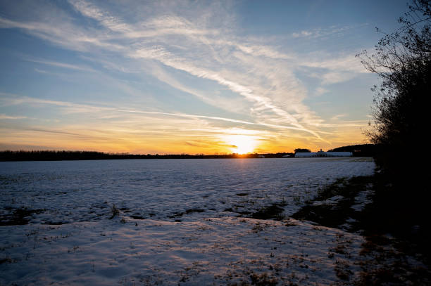 A snow-covered field at sunset with a golden sky and distant farm in Daphne, Alabama, United States