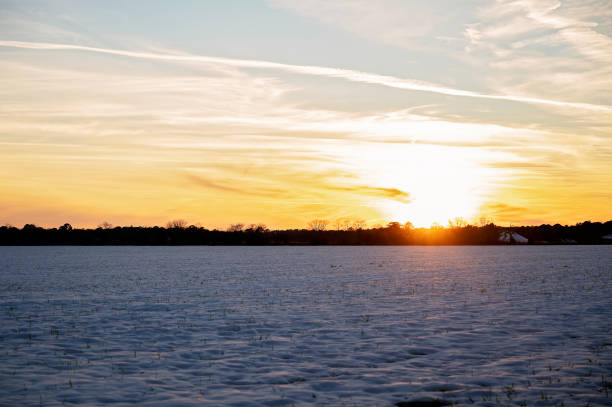 A snow-covered field at sunset with a golden sky and distant farm in Daphne, Alabama, United States