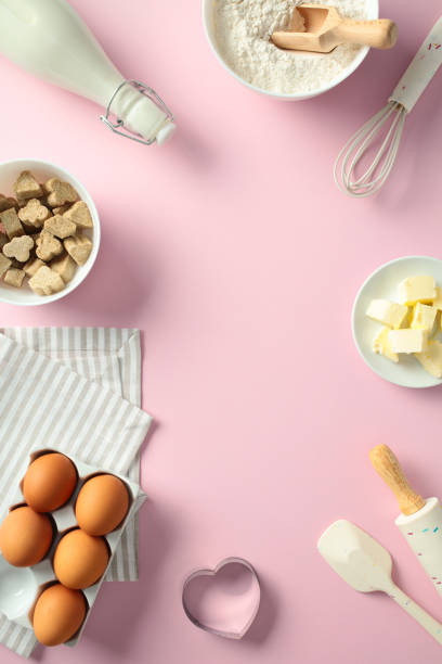 istock Baking essentials and tools on a pink background, including eggs, flour, butter, milk, sugar, a whisk, a rolling pin, and a heart-shaped cookie cutter. Flat lay, top view. 2199030915