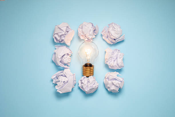 Crumpled pieces of paper are arranged around glowing light bulb against soft blue background. This visual symbolizes process of brainstorming and generating new ideas
