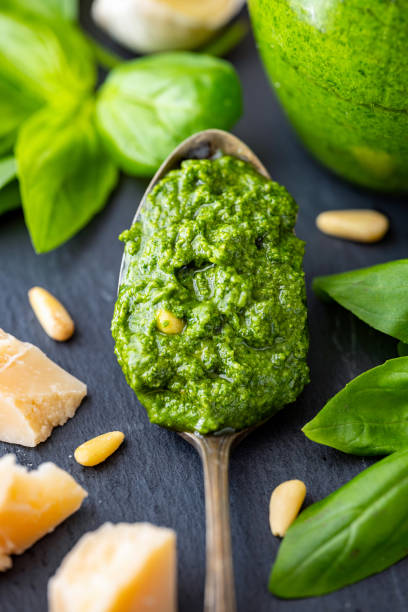 Clode-up of spoonful homemade basil pesto  sauce  with basil leaves, pine nuts, Parmesan cheese and clove of garlic in the background