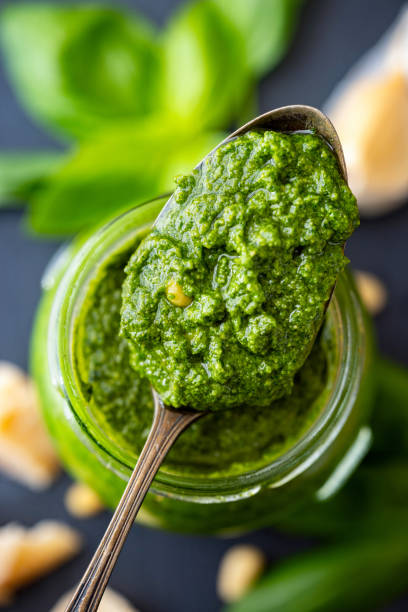 Fresh green basil pesto sauce on a spoon with basil leaves, pine nuts, Parmesan cheese and clove of garlic in the background