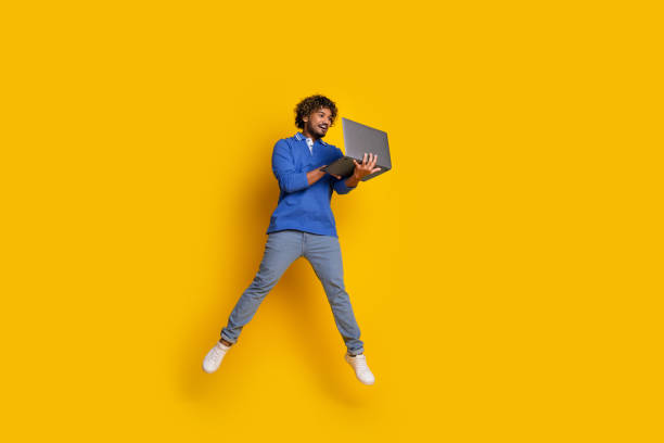 istock Happy young man jumping while holding a laptop, dressed casually in blue, surrounded by vibrant yellow background 2199049063