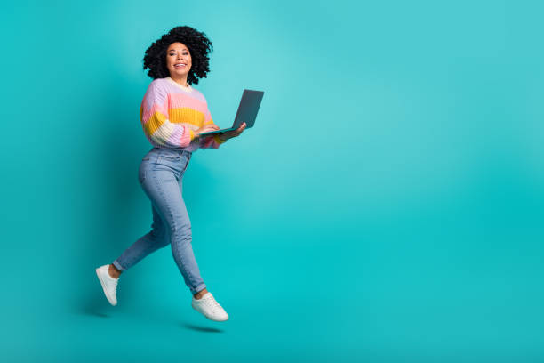 istock Energetic woman holding a laptop while jumping on a turquoise background with joyful expression 2199049594
