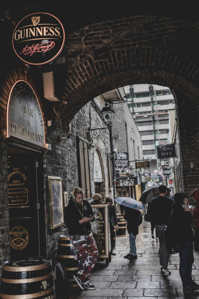 DUBLIN, IRELAND - SEPTEMBER 20, 2018: People walking under a stone arch near a pub in Dublin, Ireland, on a rainy day, with umbrellas and wet pavement.