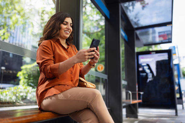 istock Smiling Woman Using Smartphone While Waiting at a Bus Stop in Urban Setting 2199054197