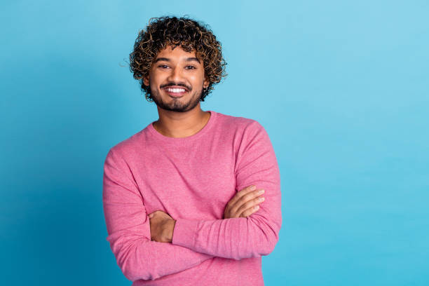 istock Confident Young Man in Stylish Pink Pullover Smiling with Arms Crossed Against a Vibrant Blue Background 2199070592
