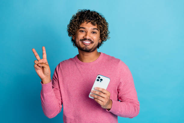 istock Young man with curly hair in pink pullover showing peace sign while holding smartphone on blue background, casual and stylish concept 2199070621