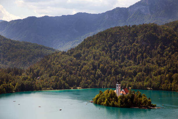 A stunning view of the picturesque Church of the Assumption on Bled Island, nestled in the middle of the emerald-green waters of Lake Bled, Slovenia. Surrounded by lush forested mountains, this iconic location offers a serene and enchanting atmosphere. The vibrant colors and tranquility make it a perfect travel destination and a photographer's dream.