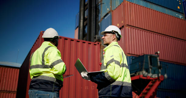 istock Two male dock managers standing and discussed over a laptop with engineer worker on the shipment in front of container box in the container warehouse area. Shipping Cargo Freights with Cargo Containers. Logistics import and export shipping yard. 2199085179