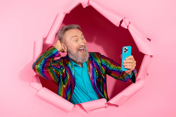 istock Cheerful elderly man in colorful disco attire, peering through torn pink paper, taking a selfie with vibrant energy 2199090474