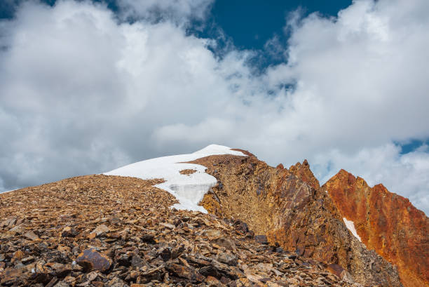 istock Snow-capped mountain ridge with cornice and red sharp rocks on top in sunlight. Snow cap and shiny pointy peak of gold color. Snow dome and colorful rocky peaked top. Sunny cloudy high mountains view. 2199107421
