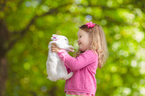 Children play with real rabbit. Laughing child at Easter egg hunt with white pet bunny. Little toddler girl playing with animal in the garden. Summer outdoor fun for kids with pets.