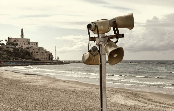 Loudspeakers or megaphones broadcasting a pole and a tsunami warning and evacuation sign located on the beach with old Jaffa in the background. Israel
