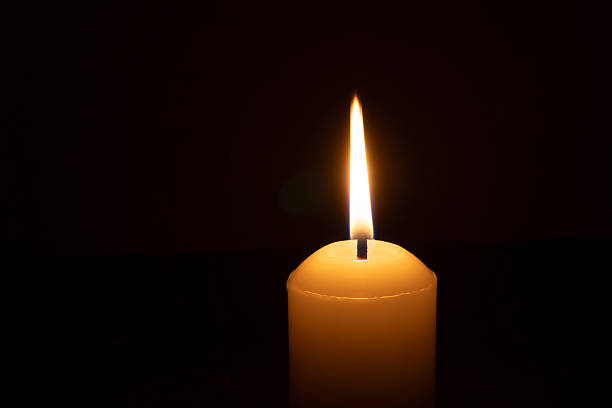 istock A single burning candle flame or light glowing on a big white candle isolated on black or dark background on table in church for Christmas, funeral or memorial service 2199115055