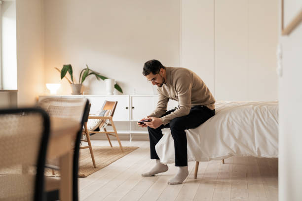istock Man sitting on bed in a cozy minimalist bedroom while using a smartphone to play online games 2199119723