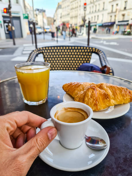 Personal perspective of a mid-adult man drinking coffee and orange juice while eating a croissant in a sidewalk café in Paris in France.