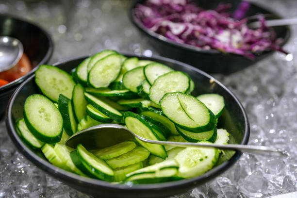 A bowl of freshly sliced cucumbers on a bed of ice, served as part of a salad bar in a buffet setting.