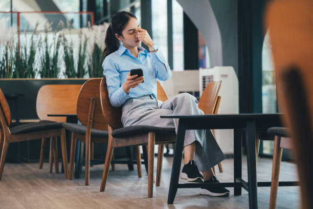Young Asian professional sitting in office chair, touching temples, displaying exhaustion while working on smartphone in contemporary workplace setting