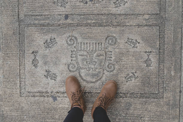 top view of brown boots standing over a painted indigenous-inspired face design on concrete sidewalk. Small pictographs resembling ancient petroglyphs.