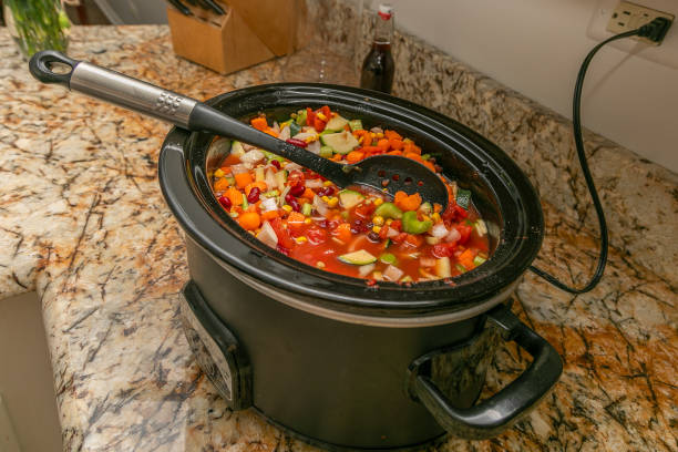 A high-angle view of vegetable soup ingredients in a crock pot and a stirring spoon. The vegetables include tomatoes, corn, carrots, zucchini, celery, onions, lentils, and kidney beans.