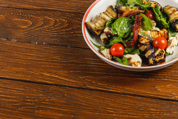 Fresh salad features grilled eggplant, spinach, cherry tomatoes, and herbs, presented in a bowl. The setting includes a textured wooden tabletop, highlighting the meal's vibrant colors.