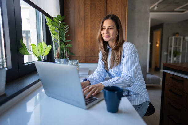 istock Young woman sitting in the kitchen, drinking coffee and using a laptop 2199145636