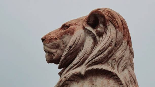 istock A weathered stone lion statue with a proud, noble expression gazes into the distance. Its detailed mane and strong features give it a commanding presence against a soft, muted sky. 2199169665
