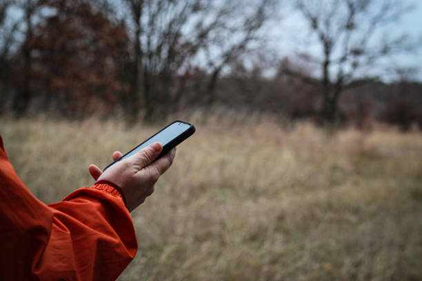 man's hand holding up a mobile phone outside isolated against a nature tree park landscape. hiking walking outdoors. active lifestyles technology and communication.