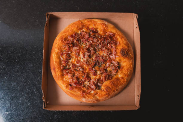 istock Top view of a ham and bacon pizza on a cardboard box ready for delivery. 2199177007