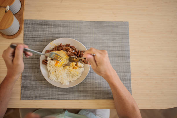 Senior Thai man enjoys a balanced lunch of rice, eggs, and meat while sitting comfortably at his dining table.