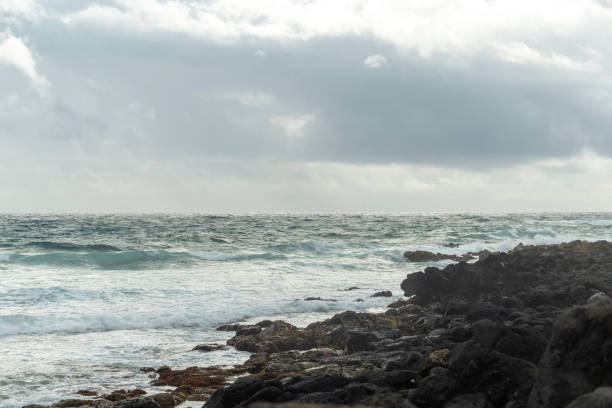 Wide shot of a woman strolling along the shoreline, taking in the dramatic waves and moody sky during her Hawaii vacation.