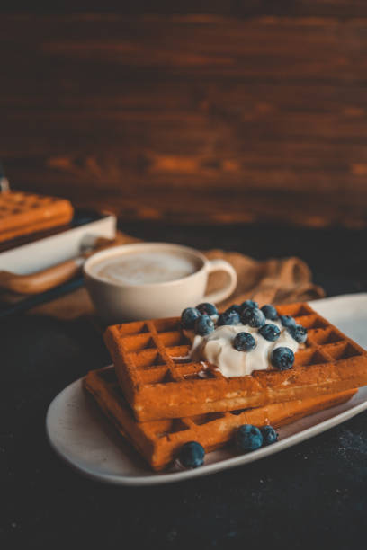 Stack of golden waffles topped with whipped cream and fresh blueberries, served on a rustic plate with a blurred cup of coffee in the background. A cozy, delicious breakfast setting.